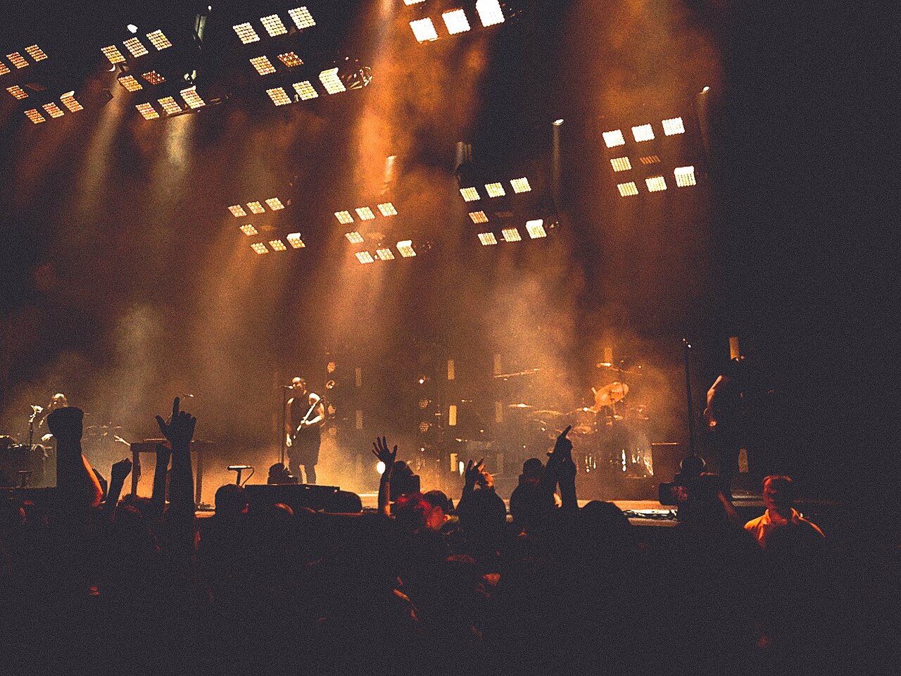 A band on a stage under rows of floodlights, seen from behind a crowd with raised hands